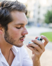 Portrait of beautiful man using asthma inhaler in park. Young man using inhaler during asthmatic attack at park. Man using a pressurized cartridge inhaler extended pharynx, Bronchodilator